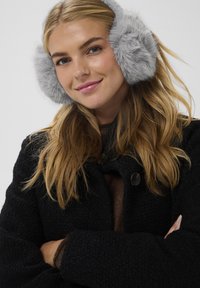 Paraorecchie in pelliccia sintetica grigia di forma circolare, con texture morbida. Indossati con un cappotto nero testurizzato e un maglione marrone, con chiusura a bottoni.