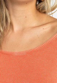 Haut orange côtelé avec un large décolleté et un tissu texturé. La couture le long du col est d'un orange plus clair contrastant.