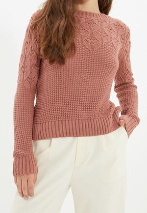 Kobieta ubrana w różowy sweter z dzianiny o chropowatej fakturze na rękawach i na piersiach, w zestawieniu z luźnymi białymi spodniami, z ręką w kieszeni.