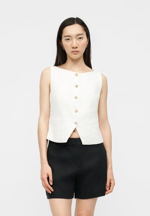 Jeune femme aux cheveux bruns et lisses porte un haut blanc sans manches boutonné et un short noir, debout devant un fond clair uni.
