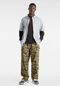 Geruite short-sleeve shirt over een zwart long-sleeve shirt; camo cargo pants met zakken; zwart-witte schoenen; staand tegen een effen achtergrond.