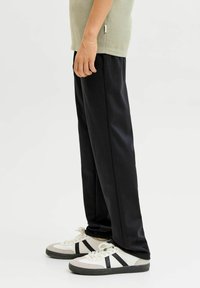 Asmuo dėvi juodus tiesius kelnes, baltus sportinius batelius su juodomis juostelėmis ir smėlinės spalvos marškinius, stovintis ant balto fono.