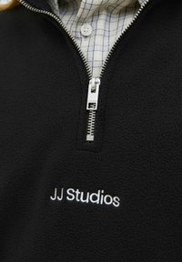 Sort fleece-pullover med sølv lynlås, med "JJ Studios" broderet i hvid. En ternet skjorte er synlig under kraven.