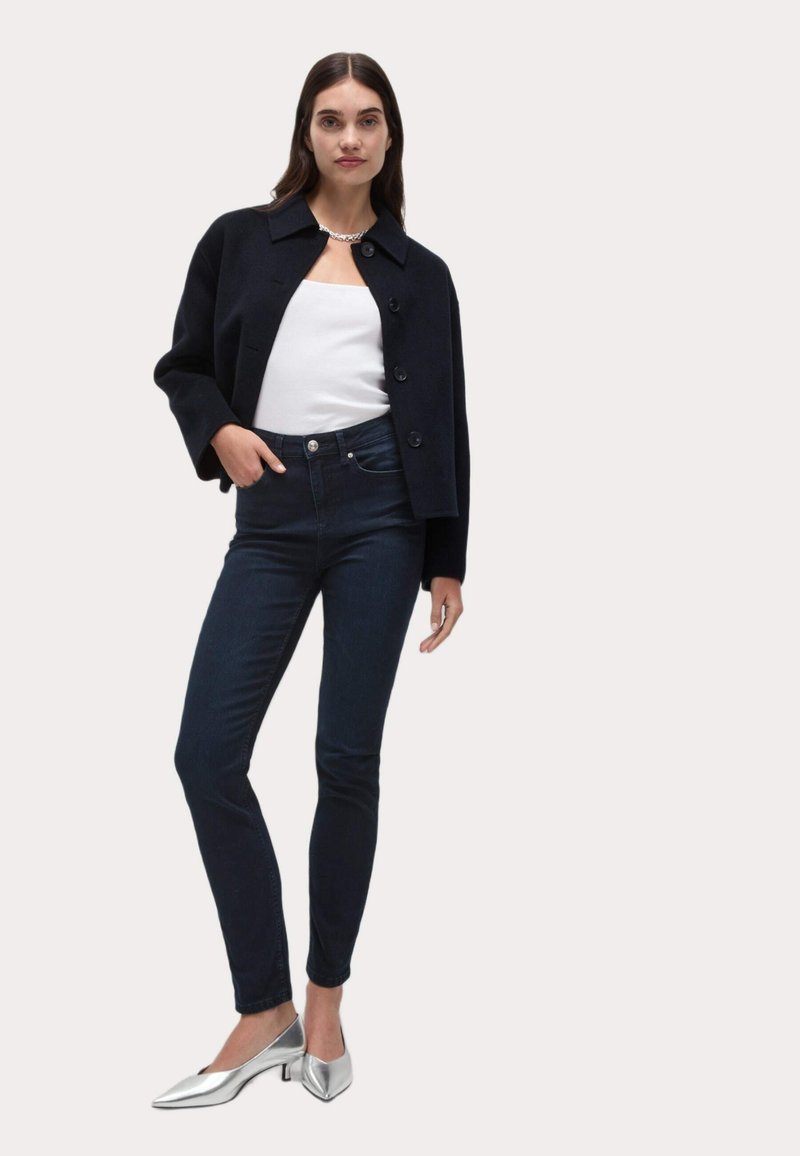 Svart kort jacka med krage och knappar, kombinerad med figursydda mörka denimjeans och silverfärgade spetsiga klackar. Släta material, minimalistisk design.