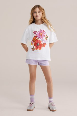 Djevojka s dugom plavom kosom nosi bijelu prezveličanu majicu s šarenim cvjetovima i tekstom "Flower Days", ljubičaste kratke hlače, prugaste čarape i srebrne cipele.