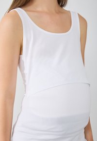 Bela materinska majica brez rokavov z okroglim izrezom. Mehka, raztegljiva bombažna tkanina z večplastnim sprednjim dizajnom, ki prilagodi rastočemu trebuščku.
