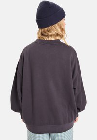 Oversized, donkergrijze sweatshirt met een ronde hals, verlaagde schouders en geribbelde manchetten. Gekleed met gemusterde groene broeken en een donkere beanie.