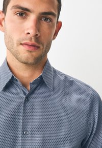 Chemise bleue à motifs avec un col boutonné, présentant un design en tissu texturé et deux boutons visibles à l'avant.