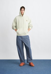 Šviesiai žaliai džemperis su priekinėmis kišenėmis ir su raišteliais, derinamas su plačiomis mėlynomis kelnėmis ir oranžiniais batais ant mėlynos grindų.