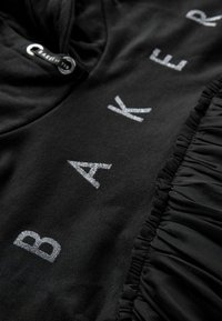 Černá mikina s kapucí vyrobená z měkké tkaniny, která má lesklé stříbrné písmena tvořící slovo "BAKER." Obsahuje detail srovnané textury na boku.