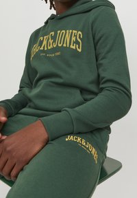 Grön sweatshirt och matchande byxor i mjukt tyg, med gul "JACK & JONES"-text och en ficka på framsidan av huvtröjan.