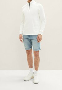 Sudadera blanca de manga larga con cremallera y textura acanalada, combinada con pantalones cortos de mezclilla azul claro. El modelo lleva zapatillas blancas y calcetines hasta el tobillo.