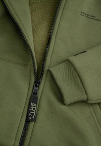 Oliwkowa bluza z kapturem z czarnym zamkiem i strukturalnym polarem wewnątrz. Posiada ściągacze przy rękawach oraz nadrukowany tekst na wewnętrznej stronie materiału.