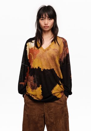 Joven con el cabello negro y lacio, lleva un suéter holgado multicolor con cuello en V y pantalones de ante marrón, con las manos en los bolsillos y expresión neutral.