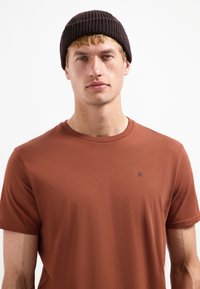 Bruine T-shirt met korte mouwen en een ronde halslijn, voorzien van een subtiele logodetail op de borst, gecombineerd met een donkere gebreide beanie.