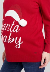 Red knit sweater featuring a white "Santa Baby" text, a white and red Santa hat design, and a fluffy pom-pom accent. Ribbed cuffs and hem.