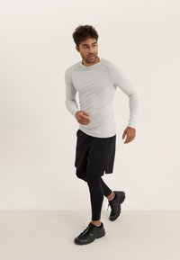 Langärmliges, hellgraues Sportshirt mit Mesh-Details, kombiniert mit schwarzen Shorts und Leggings. Schwarze Sportschuhe vervollständigen das Outfit.