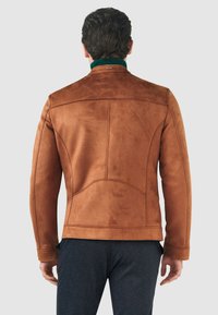 Brun mockaskinnjacka med en slät yta, upprätt krage och synliga sömnadsdetaljer. Matchad med mörka byxor. Bakifrån sedd.