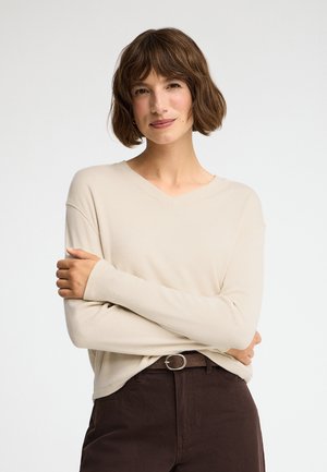 Joven mujer con cabello corto y castaño, vistiendo una camiseta de manga larga color crema y pantalones marrón oscuro, de pie con los brazos cruzados sobre un fondo claro.