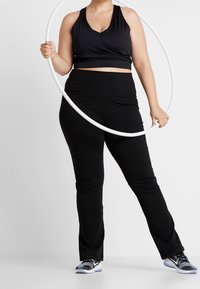 Černá sportovní podprsenka s překládaným designem, doplněná černými přiléhavými legínami. Modelka drží bílý hula hoop a má na sobě černé sportovní boty.