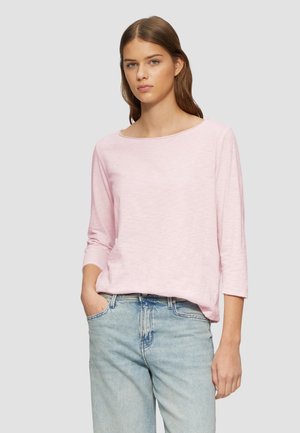 Jonge vrouw met lang bruin haar, een lichtroze shirt met driekwart mouwen en lichtblauwe spijkerbroek, met één hand in de zak.