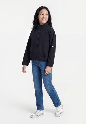 Girl with long dark hair wearing a black zip-up jacket, blue jeans, and white sneakers, smiling and looking to the side.