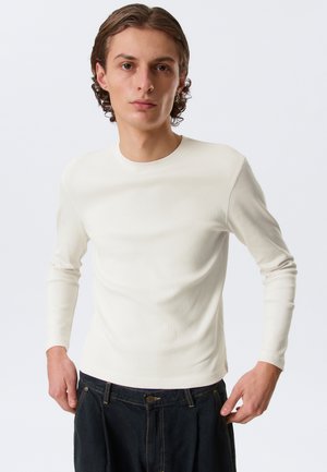 Giovane uomo con capelli corti e ricci che indossa una maglia bianca aderente a maniche lunghe e jeans scuri, in piedi contro uno sfondo chiaro e uniforme.