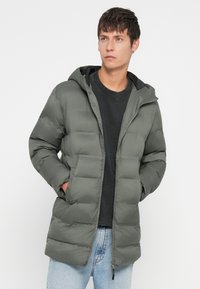 Πουφάκι σε ελιώδη πράσινο χρώμα, με κουκούλα, κλείσιμο με φερμουάρ και καπιτονέ σχέδιο. Συνδυασμένο με μαύρη μπλούζα και ανοιχτόχρωμα τζιν.