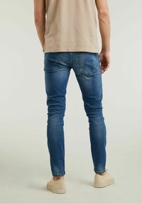 Jean slim bleu avec une texture délavée, poches arrière ornées de détails de couture discrets, associé à des baskets beige.