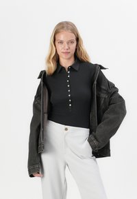 Polo shirt noir à côtes avec un bouton à l'avant, superposé sous une veste en denim foncé, associé à un pantalon gris clair.