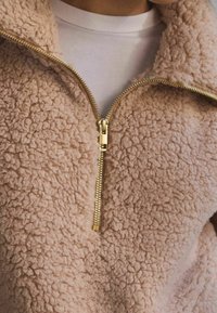 Růžový fleecový pulovr se zipem vpředu, s texturovaným povrchem, zlatými zipovými prvky a vysokým límcem. Pod ním je bílá košile.