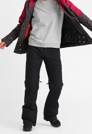 Persona con pantalones de nieve negros, una sudadera gris y una chaqueta oscura con interior rojo, de pie con los brazos ligeramente levantados.