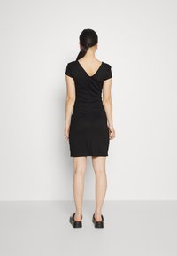 Černé přiléhavé šaty s krátkými rukávy, designem ve tvaru V na zádech, texturovaná látka, po kolena. Modelka má na sobě černé nazouvací boty a stojí před bílým pozadím.