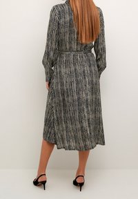 Frau mit langen, glatten Haaren, die ein tailliertes, langärmliges, schwarz-beiges gestreiftes Midi-Kleid und schwarze Absatzsandalen trägt, steht vor einer schlichten Wand.