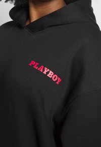 Sweat à capuche noir en coton, arborant un logo rouge "PLAYBOY" bien visible sur le côté gauche. Texture douce avec une coupe classique et une capuche.