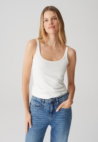 Top bianco senza maniche con scollo rotondo, abbinato a jeans blu sbiaditi. Il tessuto appare liscio, con una silhouette aderente e cuciture minime.