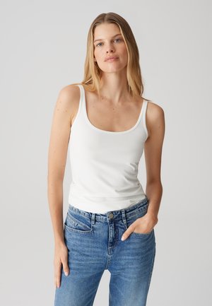 Top bianco senza maniche con scollo rotondo, abbinato a jeans blu sbiaditi. Il tessuto appare liscio, con una silhouette aderente e cuciture minime.