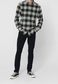 Camisa de cuadros gris y negra con cuello, mangas largas y corte relajado, combinada con pantalones oscuros y zapatillas negras.