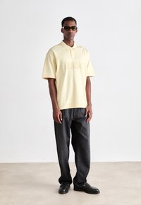 Mann trägt ein gelbes HUGO Poloshirt, schwarze lockere Jeans, schwarze Schuhe und dunkle Sonnenbrille, steht vor einer weißen Wand auf einem beigen Boden.