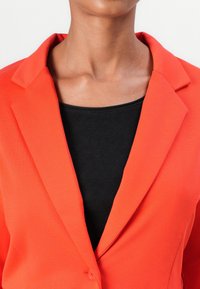 Blazer rouge avec un col cranté et une fermeture à un bouton, porté sur un haut noir. Les deux vêtements sont en tissu lisse et texturé.