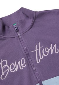 Giacca viola con zip e colletto alto, caratterizzata dal ricamo "Bene" in viola chiaro. Il tessuto è liscio, con un pannello a contrasto azzurro chiaro.