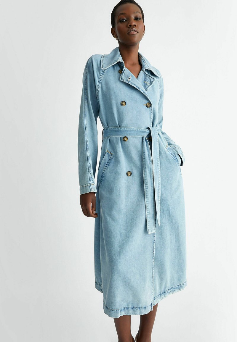 Cappotto trench in denim azzurro chiaro con design doppiopetto, ampio colletto, vita con cinturino, tasche laterali e dettagli ai polsini.