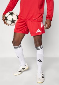 Κόκκινη αθλητική φανέλα και σορτς με λευκό λογότυπο Adidas τριών ραβδώσεων, συνδυασμένα με λευκές κάλτσες με μαύρες ρίγες και λευκά ποδοσφαιρικά παπούτσια.