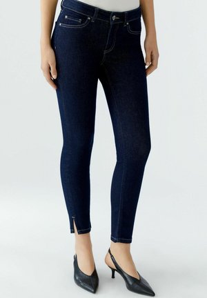 Dunkelblaue Skinny-Jeans aus Denim mit hoher Taille, ausgestattet mit einem kleinen Schlitz am Knöchel und kontrastierenden Nähten. Kombiniert mit schwarzen spitzen Heels.