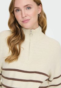 Suéter color crema con cuello alto, con rayas marrones horizontales y cierre de cremallera. Textura suave y acanalada con un ajuste relajado.