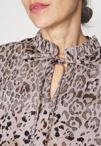 Halenka s krátkým rukávem ve světle šedé barvě s černým a hnědým leopardím vzorem. Má volánkový výstřih a zavazování na mašličku vpředu.