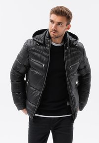 Chaqueta acolchada negra con capucha, que presenta bolsillos con cremallera, textura suave, diseño acolchado y acentos de hardware plateado.