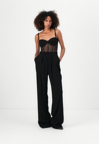 Schwarzes Zweiteiler-Outfit mit einem durchsichtigen, taillierten Oberteil und hochgeschnittenen, weiten Hosen. Betont durch einen mittigen Reißverschluss und schlichte Linien.
