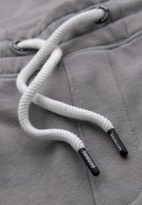 Zbliżenie na biały sznurek ze czarnymi końcówkami oznaczonymi napisem "DIMBRE" na szarym materiale, prawdopodobnie część pasa bluzy z kapturem lub spodni dresowych.