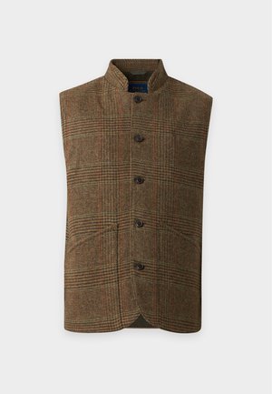 Homme debout les mains dans les poches portant une chemise rouge, un gilet marron boutonné, un pantalon blanc et des mocassins beige sur un fond uni.